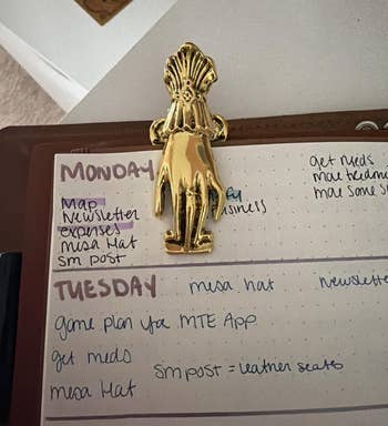 Gold hand-shaped clip holding a planner with handwritten weekly tasks, including meetings and newsletter tasks