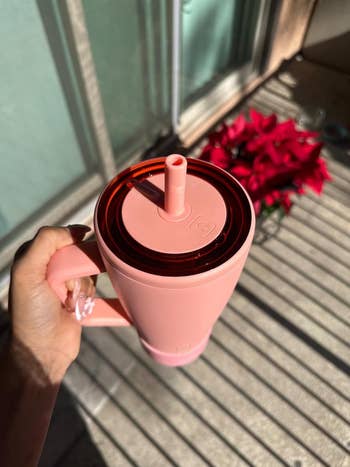 A hand holding a reusable pink tumbler with a straw, next to a potted plant on a sunlit balcony