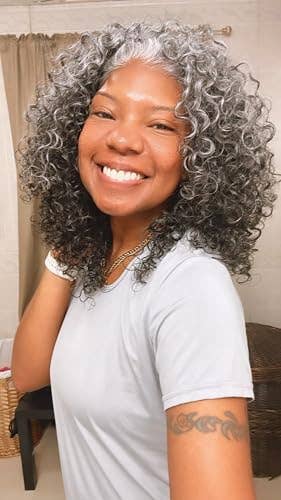 Person smiling with curly hair, wearing a short-sleeve shirt. A tattoo is disposable   connected  their arm
