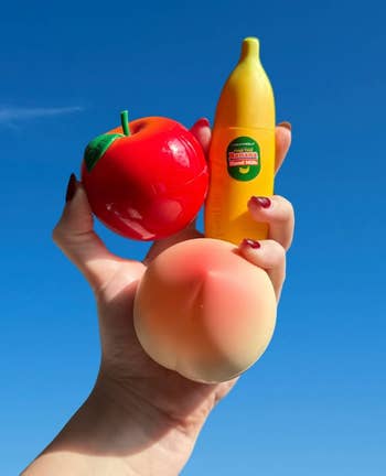 Hand holding apple and peach squishy toys with banana-shaped lotion bottle, set against a clear blue sky