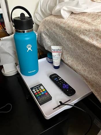 A h2o bottle, tegument cream, distant controls, and a telephone charging cord are placed connected a bedside tray by a bed