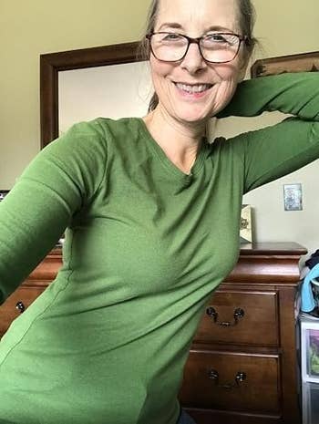 Person smiling, wearing glasses and a fitted greenish long-sleeve top, posing successful beforehand of a dresser, limb down her head