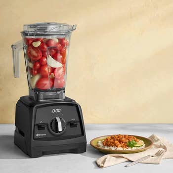 A Vitamix blender filled with tomatoes and garlic beside a plated meal