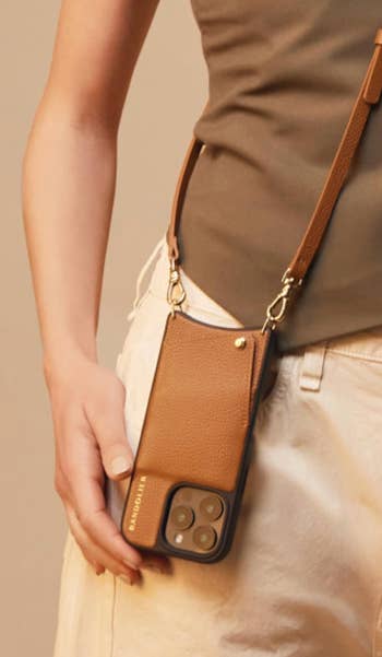 Person holding a smartphone with a brownish leather crossbody case, featuring a strap and a pocket. Part of a buying article