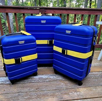Three blue suitcases with yellow straps are displayed on a wooden deck, possibly for a shopping review