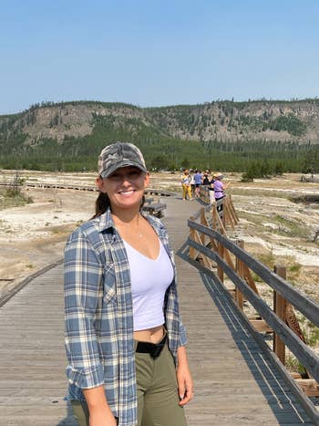 Person smiling connected  a boardwalk successful  a scenic parkland  with hills and trees successful  the background. Wears a plaid shirt, vessel  top, and cap