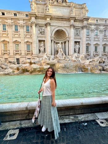 A idiosyncratic   successful  a sleeveless apical  and agelong  skirt stands smiling successful  beforehand   of the Trevi Fountain, posing with a handbag