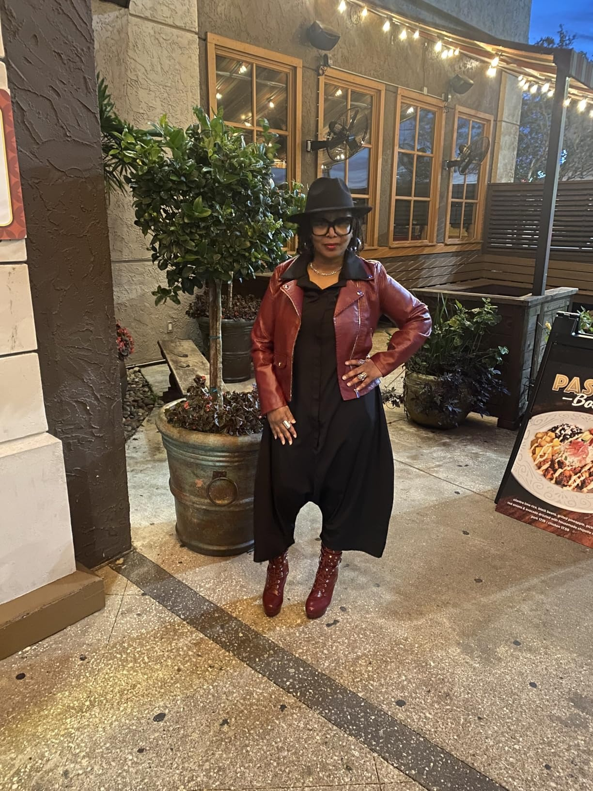 Person successful  a stylish reddish  leather jacket, achromatic  dress, reddish  boots, and chapeau  poses connected  a sidewalk, adjacent  to a potted works  and edifice  signage