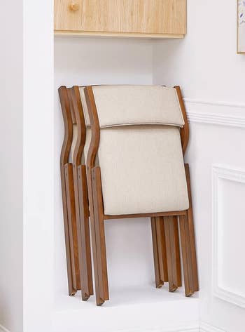 Beige folding chair with wood body, leaned against white wall underneath a shelf