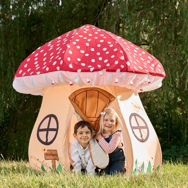 Dua anak tersenyum dan mengintip dari dalam tenda bermain berbentuk jamur dengan desain jendela, di luar ruangan.  Tanda “Selamat Datang” tergantung di pintu