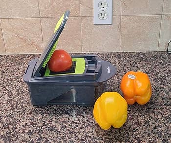 Kitchen countertop with a food chopper holding a tomato, next to a yellow and an orange bell pepper