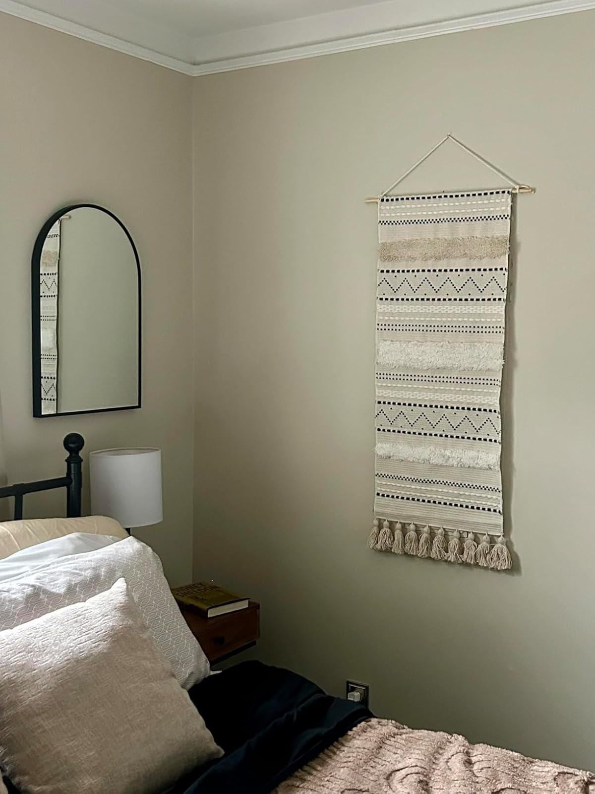 Bedroom with wall tapestry, black metal bed frame, round mirror, and nightstand with lamp. Neutral decor creates a cozy, minimalist look