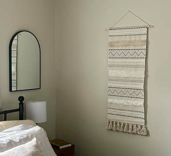 Bedroom with wall tapestry, black metal bed frame, round mirror, and nightstand with lamp. Neutral decor creates a cozy, minimalist look