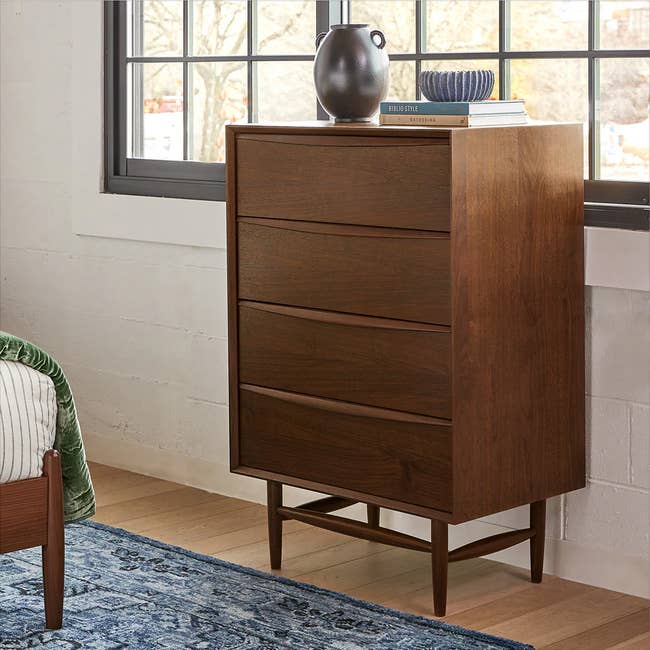 A mid-century modern wooden dresser with four drawers, a vase on top, next to a window
