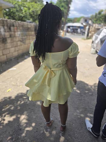 A person outdoors facing away, wearing an off-shoulder, knee-length yellow dress with a bow at the back, and white sandals