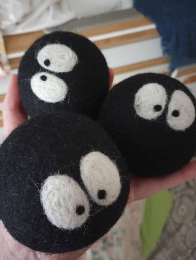 Hand holding three black wool dryer balls with cute white, animated eyes. Suitable for laundry use and eco-friendly shopping options