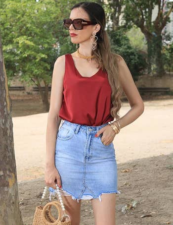 Woman in sleeveless top and denim skirt holding a woven bag, accessorized with large sunglasses and statement earrings, outdoors