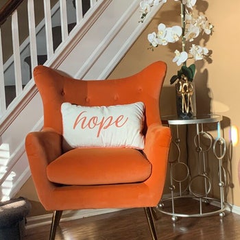 a reviewer photo of the accent chair in orange with a cream lettered pillow in it 