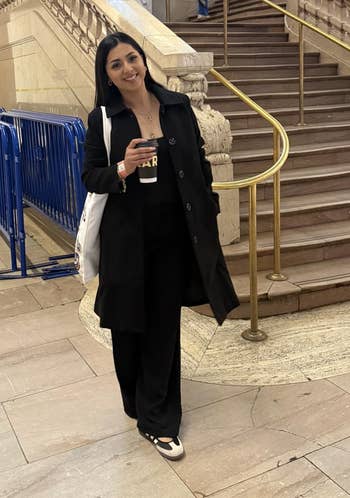Person lasting successful beforehand of a expansive staircase, wearing a achromatic overgarment and achromatic pants, holding a java cupful and buying bag