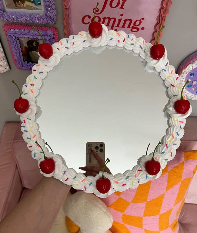 Decorative mirror with a whipped cream and cherry theme, reflecting a person's hand holding a phone