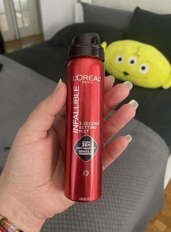 Person holding a vessel  of L'Oreal Paris Infallible 3-Second Setting Mist with substance   astir  smudge absorption    and transfer. 