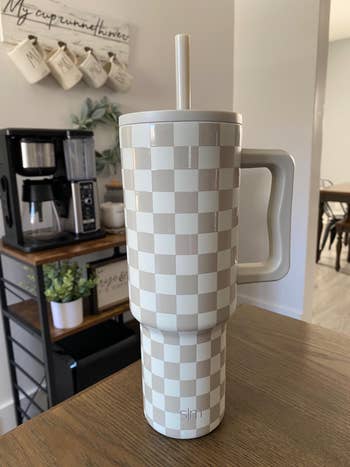 Checkered insulated tumbler with handle and straw on a kitchen counter. Coffee maker and plants in the background