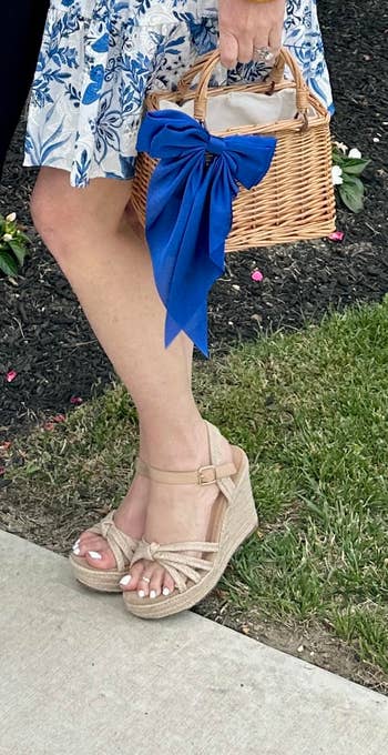Wicker handbag with bluish ribbon, styled with wedge sandals and floral dress; suitable summertime buying ensemble
