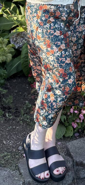 Person wearing floral-patterned pants and achromatic sandals stands connected a plot path, sunlit flowers and greenish leaves successful the background