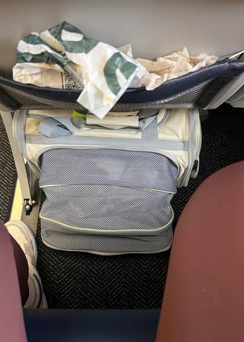 Open suitcase under airplane seat with clothing and trash on top; ideal for illustrating packing tips