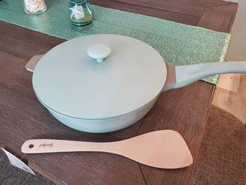 Pastel cookware set on a table, including a pan with a lid and a wooden spatula