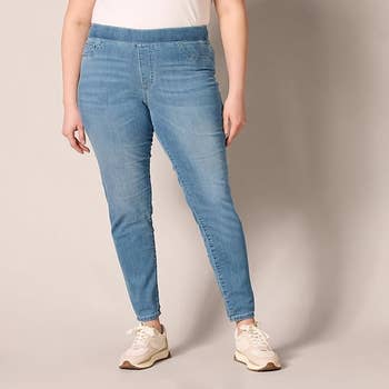 Orang yang memodelkan jegging denim biru dan sepatu kets putih, berdiri dengan latar belakang netral. Pakaian santai untuk pakaian sehari -hari
