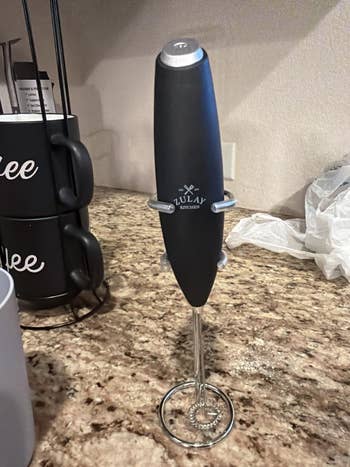 Electric beverage frother with a basal connected a room countertop, adjacent to stacked java mugs