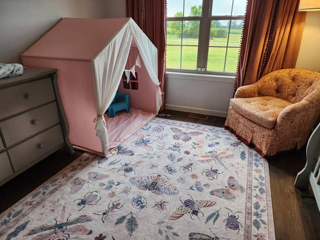 Children's room with a play tent, dresser, floral rug, and upholstered chair by a window overlooking a grassy field