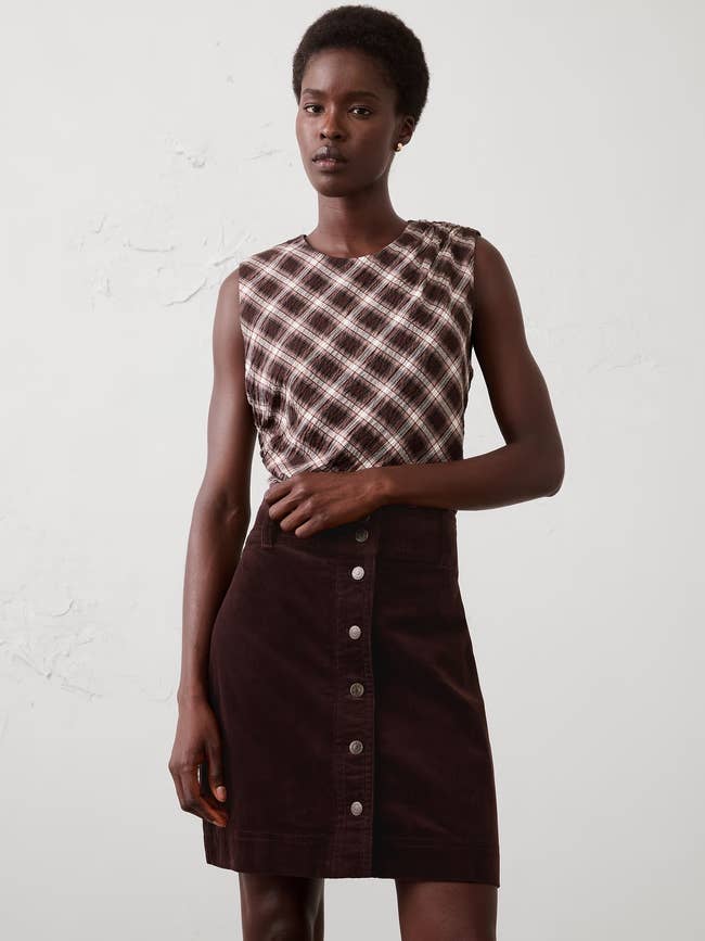 A idiosyncratic   models a sleeveless plaid apical  paired with a buttoned, knee-length skirt