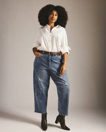 Person wearing a achromatic button-up shirt, relaxed jeans, and ankle boots lasting confidently against a plain background