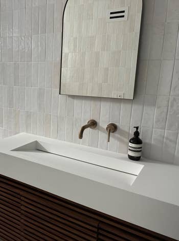 Modern bath with wall-mounted sink, sleek faucet, shower, and toilet. Minimalist plan with tile walls and woody furniture accents
