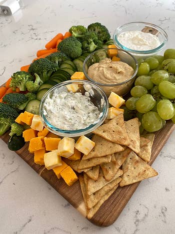 A food platter with broccoli, cucumbers, carrots, grapes, cheese cubes, crackers, and three dips: hummus, ranch, and a white dip