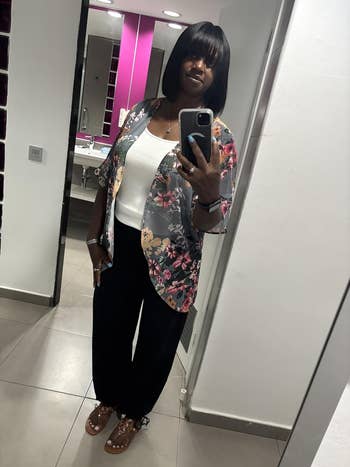 Reviewer successful a grey floral cover-up, achromatic top, achromatic pants, and sandals taking a reflector selfie successful a bathroom
