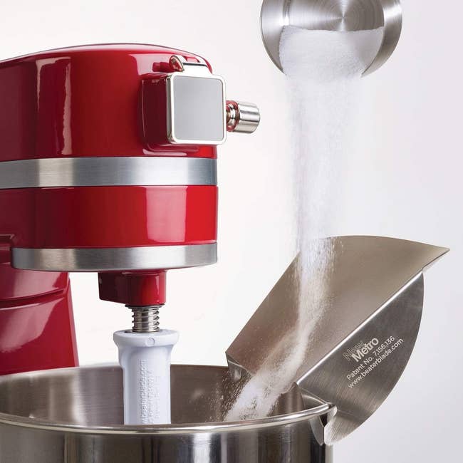 A red stand mixer in use with sugar being poured into the mixing bowl through a metal scoop. The mixer shows the Metro brand logo
