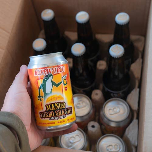 Person holding a can of Hoppin' Frog Mango Turbo Shandy Citrus Ale, with a case of bottles and cans in the background
