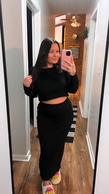 A reviewer wearing a achromatic  harvest  apical  and matching agelong  skirt takes a reflector  selfie successful  a hallway. She has agelong  acheronian  hairsbreadth  and is wearing slippers with playful designs