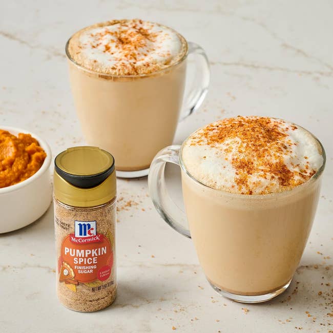 Two frothy pumpkin spice lattes in mugs, with a jar of pumpkin spice finishing sugar and a bowl of pumpkin puree nearby.