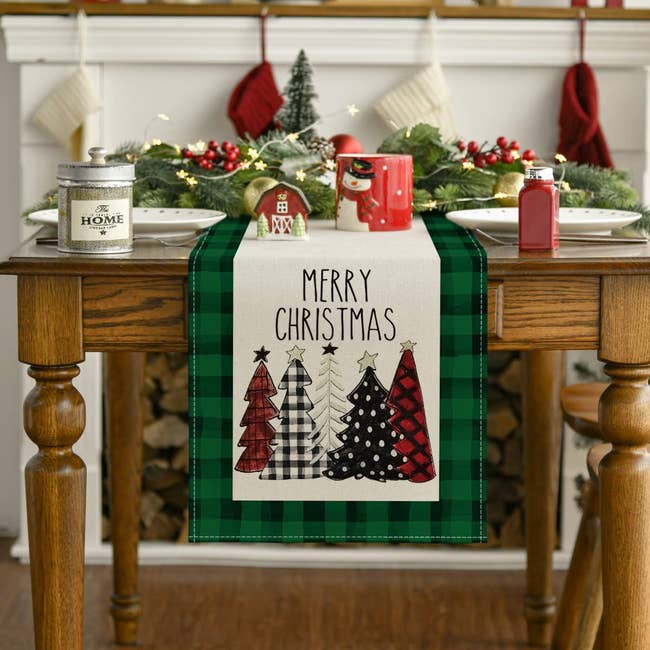 a green and black buffalo check edges table runner with white in the middle and five trees on the end with merry christmas above them
