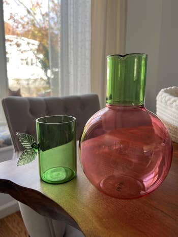 Glass pitcher and tumbler connected a woody table. The pitcher is pinkish with a circular shape, and the tumbler is greenish with leafage detail, suitable for decor