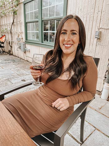 Person successful a fitted brownish formal sits outdoors, smiling and holding a solid of reddish vino astatine a patio table