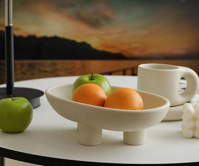 A cream, three-legged ceramic bowl holding fruit
