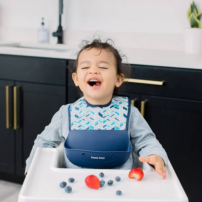 Toddler successful a highchair, wearing a patterned bib, smiles with scattered blueberries and strawberries connected the tray