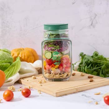 Salad jar with layers of veggies, nuts, and grains connected  a woody  board, surrounded by greens and vegetables, suggesting a fresh, steadfast   purchase