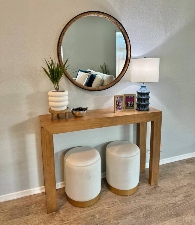 Round mirror above a console table with decorative items and two ottomans underneath. Perfect for a home entryway