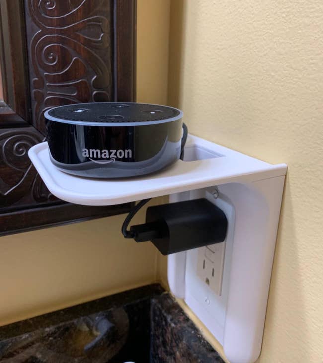 A white small shelf attachment installed on an outlet with a plugged in Amazon Alexa on top 